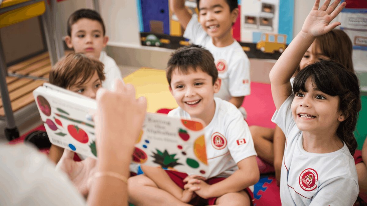 Crianças felizes do ensino infantil da MapleBear respondendo perguntas de ingles a professora que esta com livro na mão.