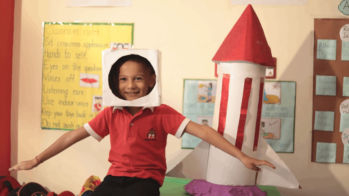Aluno da educação infantil sentado e brincando de astronauta em frente a um foguete de papel na MapleBear.