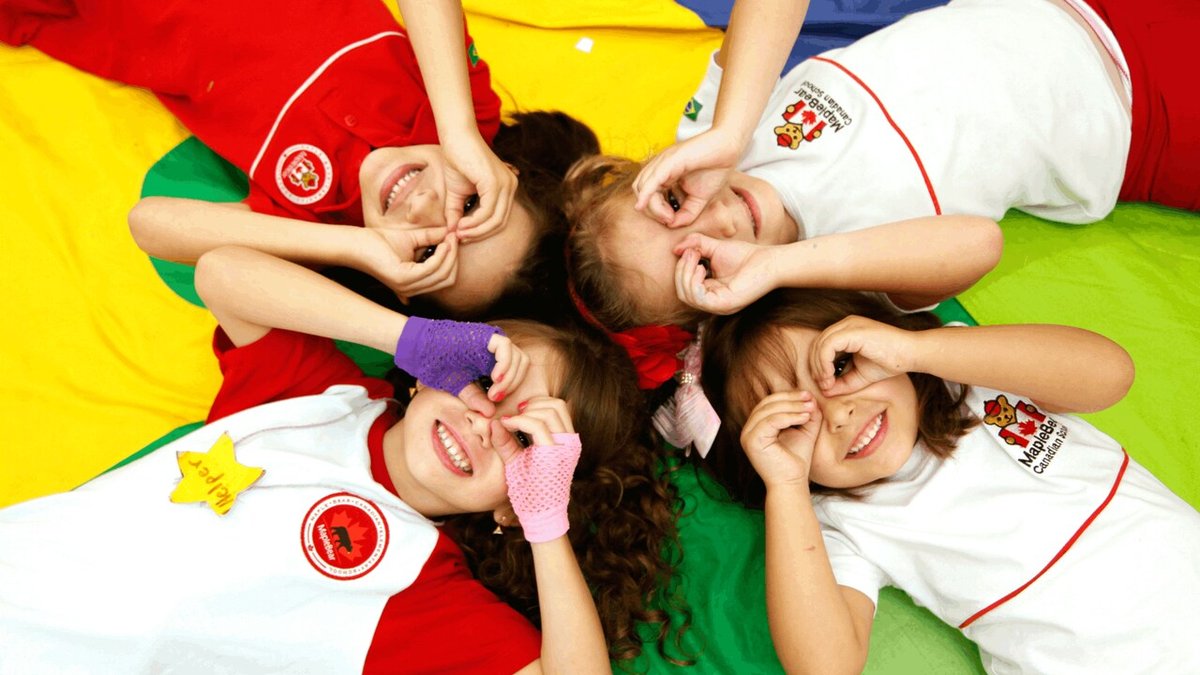 Quatro crianças felizes, vistas de cima, deitadas no chao sobre a bandeira do Brasil, usando uniforme da MapleBear.